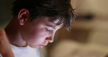 Samimi genç bir çocuk gece ekrana bakıyor. Işık saçan tablete bakan çocuk yüzü