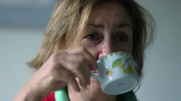Senior woman holding cup of coffee in the morning breakfast table, close-up face of elderly lady sips warm drink while interacting with people off-camera