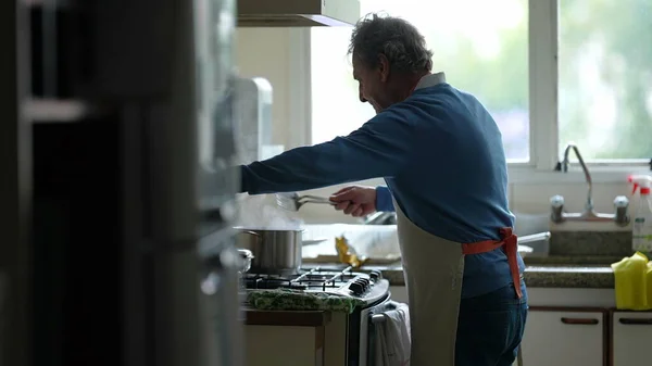 Candid senior man cooking food standing by kitchen stove stirring ingredients with utensil. Elderly retired person preparing meal wearing apron, authentic scene