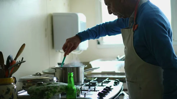 Senior man sipping beer from bottle while cooking food by kitchen stove, apron wearing older person drinks alcoholic beverage while preparing meal