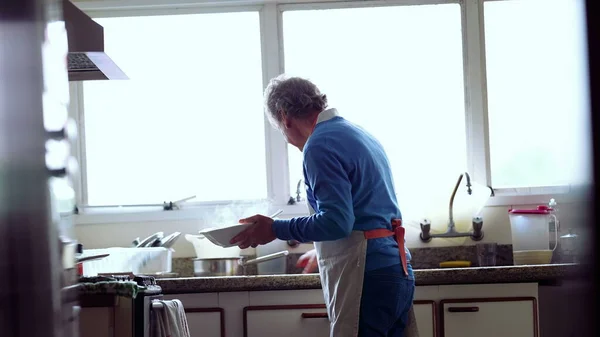 Elderly Chef in Apron Cooking by Home Kitchen Sink, candid and authentic of retired person preparing meal
