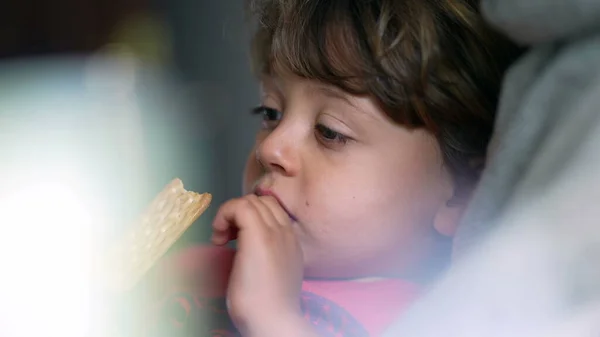 Pensive child eating cookie while laid down on couch, close-up face of small boy snacking sweet treat while in contemplation