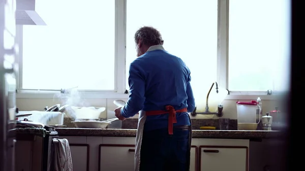 Back of senior chef preparing meal at home by kitchen sink. Retired older man cooking food, wearing apron. casual domestic lifestyle in old age