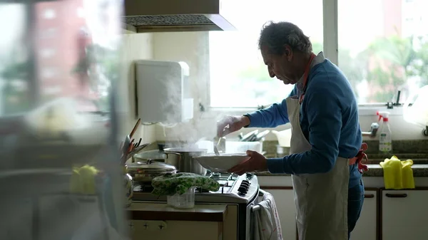 Candid senior man cooking food by kitchen stove, elderly person preparing meal for family, wearing apron, authentic retirement lifestyle