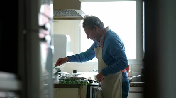 Retired Chef in Apron Focused on Home Cooking, candid senior man preparing food by kitchen stove