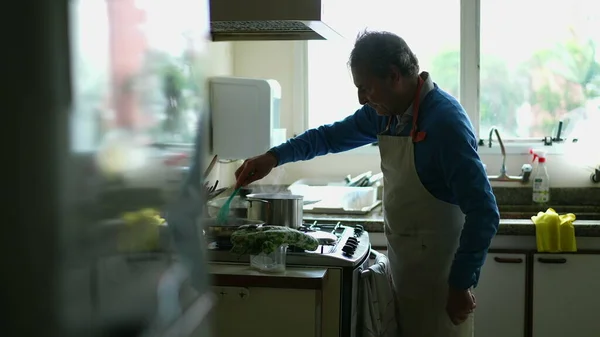 Candid senior man cooking food by kitchen stove wearing apron. One elderly caucasian person preparing meal for family