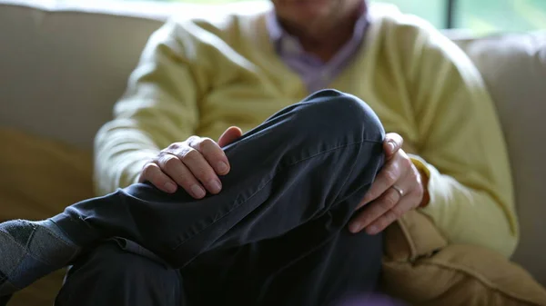 Senior man with legs crossed, detail close-up of older person seated on couch sofa at home wearing yellow sweater and pants