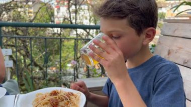 Young boy at lunch table with family outside in home patio. Child eating pasta and drinking juice