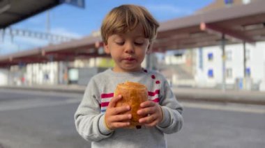 One cute little boy snacking chocolate croissant bread standing at train station platform. Child eating carb food. Pain au chocolat.