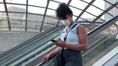 Commuter woman wearing face mask at moving escalator checking smartphone device. Person entering subway with mask prevention.