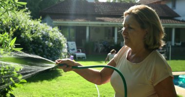 Older woman gardening and water plants with water hose outside in sunlight