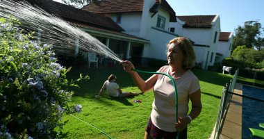Happy older woman in 60s watering garden with water hose