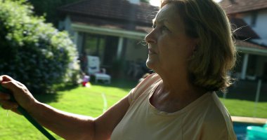 Woman in 60s watering plants with water hose outdoors with sunlight lens-flare