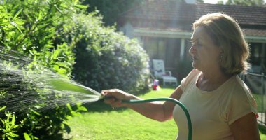 Older woman gardening and water plants with water hose outside in sunlight