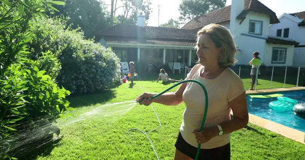 Senior woman watering plants with water hose outside. Relaxed older lady in 60s taking care of garden
