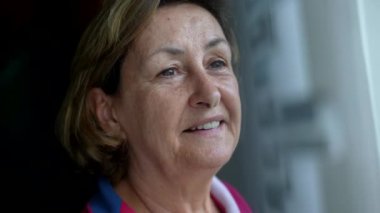 Yakın plan mutlu, düşünceli yaşlı kadın pencerenin kenarında durmuş manzaraya bakıyor ve sessiz bir sevinç hissediyor. Emekli Kafkasyalı yaşlı bayanın düşünceli ifadesi.