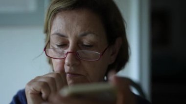 Yaşlı kadın akıllı telefon cihazıyla internetten içeriği okurken burnunu ve yüzünü kaşır. Konsantre Kafkasyalı kişi