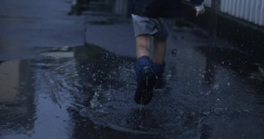 Çocuk sevinçle yağmur botlarıyla su birikintisine koşuyor.