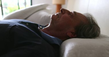 Older man lies in bed to rest, senior looks at camera while laying down