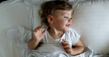 Cute baby lying in bed covering face with hands, adorable infant toddler playing