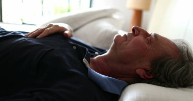 Older man lies in bed to rest, senior looks at camera while laying down