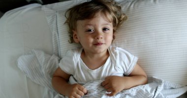 Cute baby lying in bed covering face with hands, adorable infant toddler playing