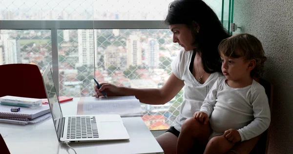 Mother multi-tasking, holding baby infant and studying from home. Candid authentic and real life mom working and parenting