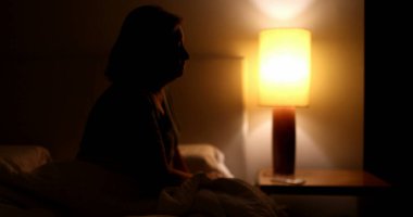 Woman suffering from insomnia gets out of bed and sits on the side unable to sleep, turns night stand light