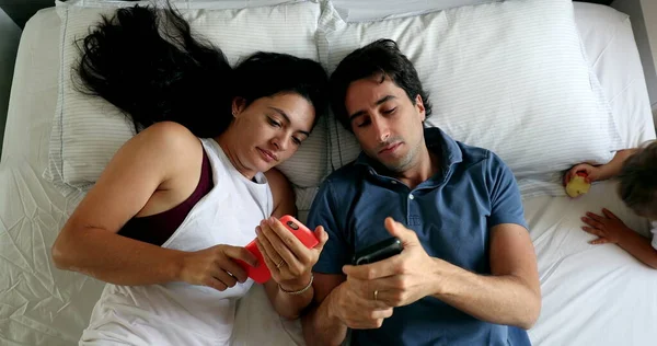 Casual couple in bed lying down looking at their cellphones, top view