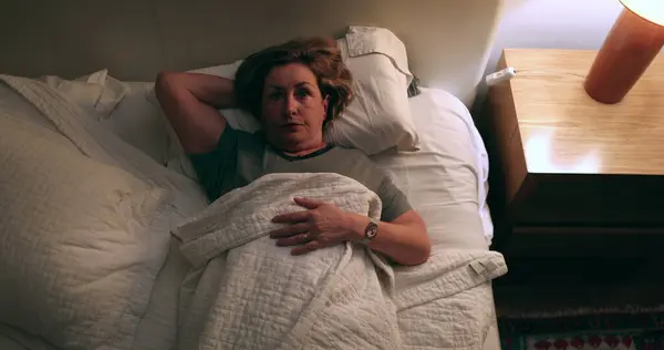 Pensive older woman lying down in bed, turns off nightstand light off and goes to sleep