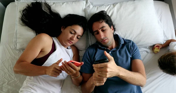 Casual couple in bed lying down looking at their cellphones, top view