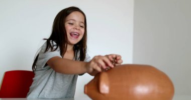 Small girl saving money, adding coins inside piggy bank