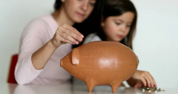 Mother and daughter financial savings adding coins inside piggy bank