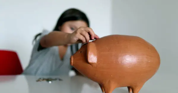 Small girl saving money, adding coins inside piggy bank