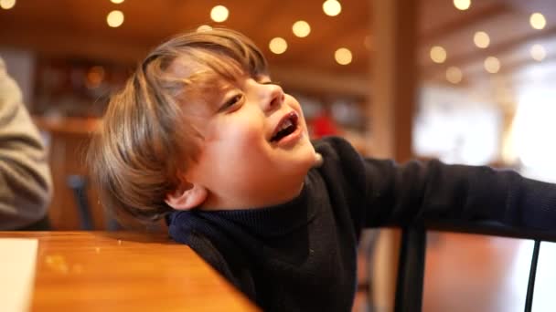 一个小男孩坐在餐馆里等待着食物的到来 他感到不耐烦 无所事事 但又带着微笑和快乐 — 图库视频影像