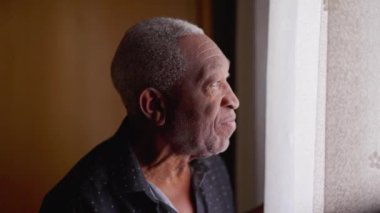 Düşünceli bir siyah Brezilyalı kıdemli adam pencerenin yanında durup düşünceli bir ifadeyle dışarı bakıyor. Afro-Amerikalı, yakından profil yüzü. Evden dışarı bakıyor.