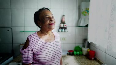 Düşünceli bir Afrikalı Amerikalı son sınıf öğrencisi mutfak penceresinin yanında durup düşünceli bir ifadeyle dışarı bakıyor.