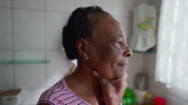 Mutfak penceresinin yanında derin bir yansımayla kıdemli siyahi bir kadın duruyor. El pençe divan duran düşünceli Afrikalı Amerikalı yaşlılar.