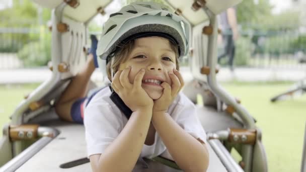 公園の楽しみ 金属の遊び装置で笑うヘルメットの男の子は 閉じたズームアウトで手のチンを休ませます — ストック動画