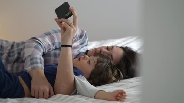 穿着睡衣的松懈的母子 在床上共享一个看在线内容的糖果时刻 — 图库视频影像
