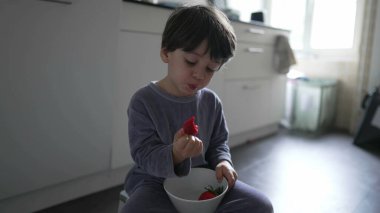 Çocuk mutfakta yerde pijama giyerek çilek yiyor. Küçük çocuk sabah kahvaltısını elinde kaseyle yiyor ve meyve tadıyor.