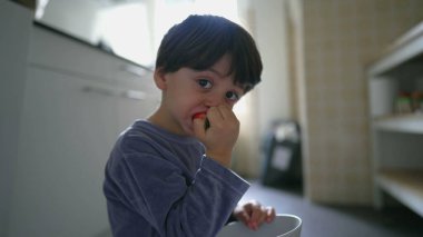 Pijamalı küçük çocuk kahvaltıda çilek yiyor, mutfakta elinde kaseyle yerde oturuyor.