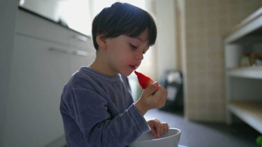 Pijamalı küçük çocuk kahvaltıda çilek yiyor, mutfakta elinde kaseyle yerde oturuyor.
