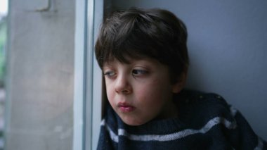 Düşünceli, düşünceli, sıkılmış bir ifadeyle evden dışarı bakan üzgün çocuk. Yakın plan küçük çocuk konutuna kapatıldı.