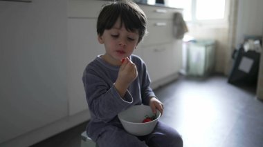 Çocuk mutfakta yerde pijama giyerek çilek yiyor. Küçük çocuk sabah kahvaltısını elinde kaseyle yiyor ve meyve tadıyor.