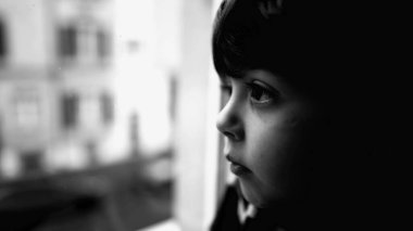 Üzgün, düşünceli, yakın plan bir çocuk. Dramatik siyah beyaz, yalnız, üzgün küçük bir çocuk gibi pencereden dışarı bakıyor.