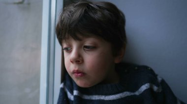 Düşünceli, düşünceli, sıkılmış bir ifadeyle evden dışarı bakan üzgün çocuk. Yakın plan küçük çocuk konutuna kapatıldı.