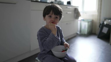 Çocuk mutfakta yerde pijama giyerek çilek yiyor. Küçük çocuk sabah kahvaltısını elinde kaseyle yiyor ve meyve tadıyor.