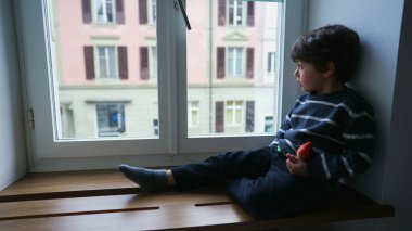 Düşünceli küçük çocuk pencerenin kenarında oturmuş pencereden dışarı bakıyor ve çiseleyen yağmura bakıyor. Sonbahar sezonunda sıkılan düşünceli çocuk ikinci kattan içeri bakıyor.