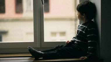 Melankolik küçük çocuk pencerenin kenarında oturmuş evden çiselemeye bakıyor, süveter giyen çocuk derin düşüncelere dalmış bir şekilde manzaraya bakıyor.